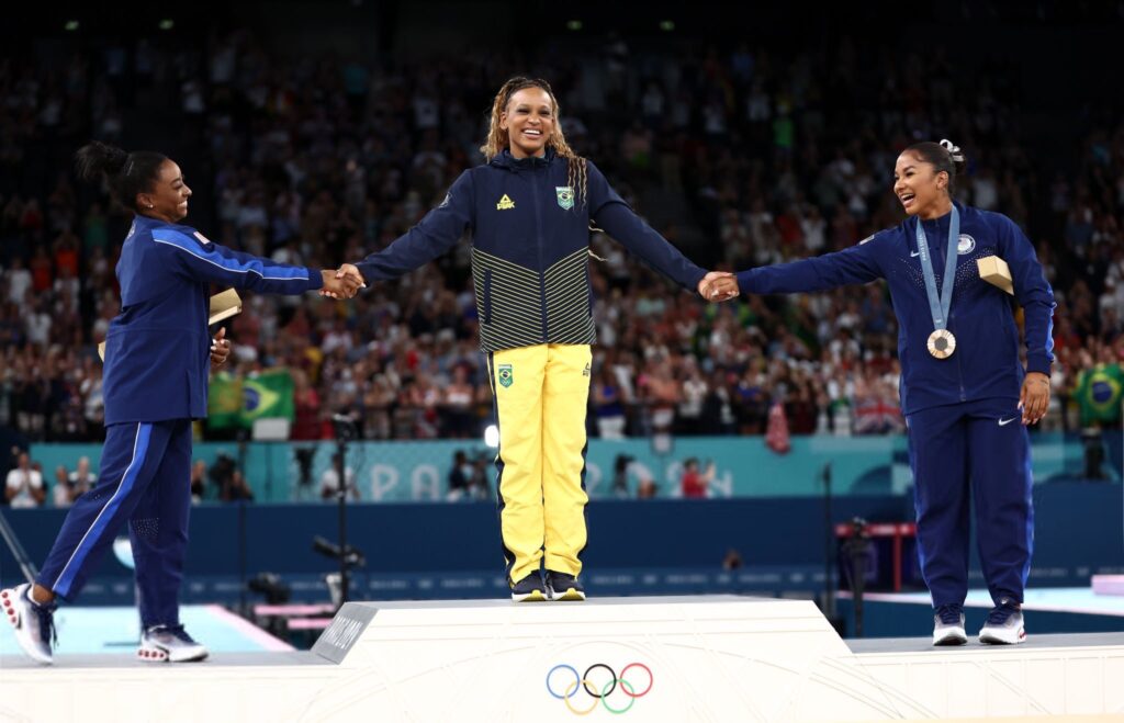 Simone Biles, Rebeca Andrade, and Jordan Chiles in the gymnastics venue at the Paris Olympics holding hands during the Floor Exercise Event Final victory ceremony