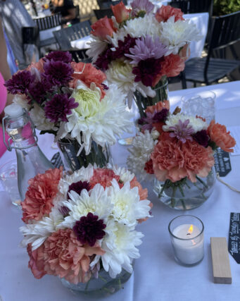 Small bouquets of various flowers in short glass vases on a a table with a white table cloth and a votive candle.