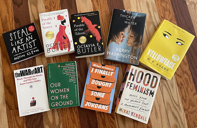 Books on a multicolor wood floor, clockwise: Steal Like an Artist, Parable of the Sower, Parable of the Talents, Thicker Than Water, Yellowface, Hood Feminism, I Finally Bought Some Jordans, Our Women on the Ground, The War of Art.