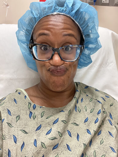 Author seated in a hospital gown, blue surgery cap, face scrunched up in a silly way.