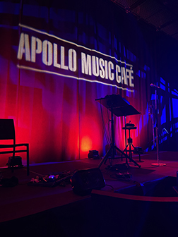 Stage with red, blue and purple lights. "Apollo Music Cafe" in white lights projected onto the curtain background. Music stand and mic onstage.