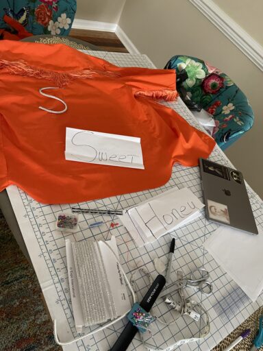 Table covered in white cardboard sewing cutting board, with measuring tape, shears, pins, a sharpie, white chalk, a seam ripper, a MacBook with stickers, rhinestone trim cording, an orange shirt with orange fringe, pieces of paper that have "sweet" and "honey" written on them.