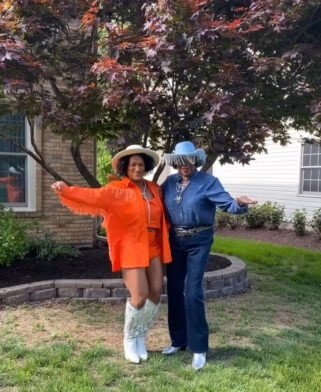 A woman in orange and a woman in denim stand together in front of a Japanese maple tree.