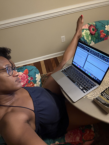Author sitting at a table with a laptop and book. One leg propped up on the adjacent chair.