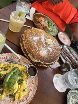 Table with plates full of pancakes, scrambled eggs with avocado, eggs with red and green salsa, and two margaritas.