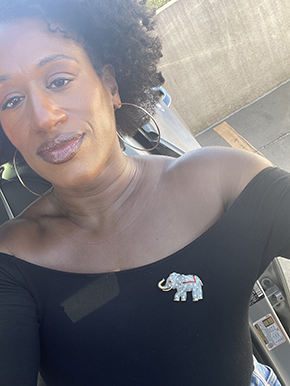 Woman in a black off shoulder top wearing an elephant pin and hoop earrings. Sitting in the car with the door open.