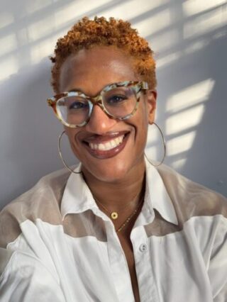 Black woman in a white shirt, her hair is short, natural and a bronze color. She is also wearing turquoise and brown glasses.