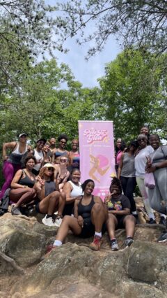 Group of women seated around the PoleToGlow banner at the summit of their mountain hike.